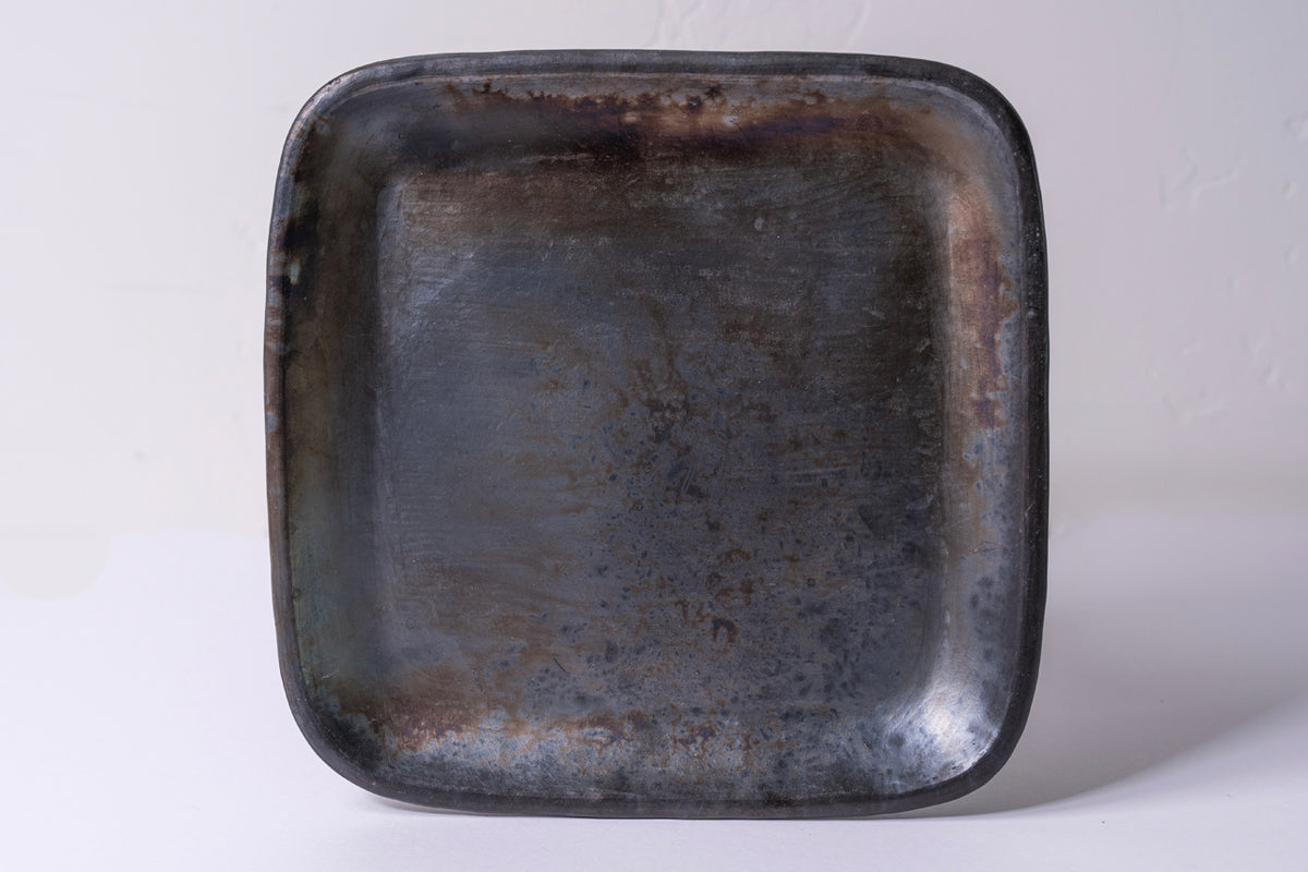 Dark gray-black handmade ceramic plate with rounded square shape and unglazed textured surface.