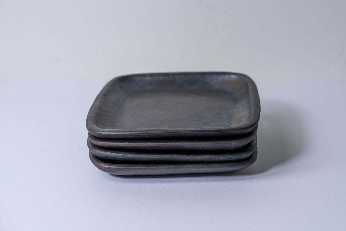 Stack of four dark gray square pottery plates with shallow wells and rounded corners.