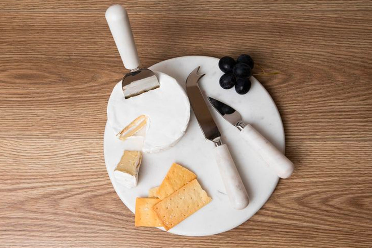White marble cheese board with three stainless steel tools and handles, displayed with cheese and grapes.