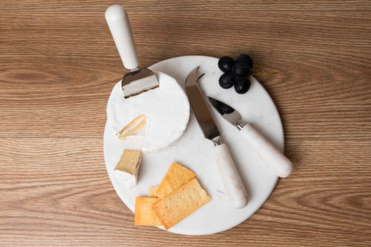 White marble cheese board with three stainless steel tools and handles, displayed with cheese and grapes.