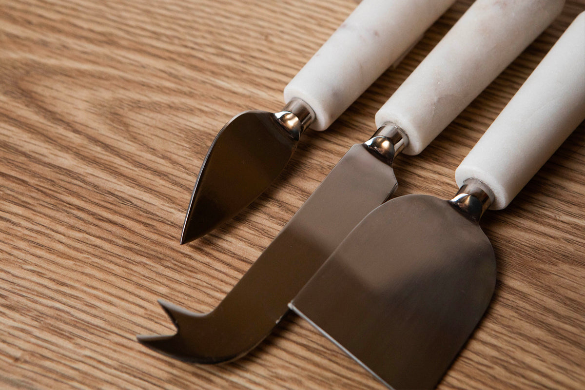 Three cheese tools with white marble handles and stainless steel blades arranged on wood surface.
