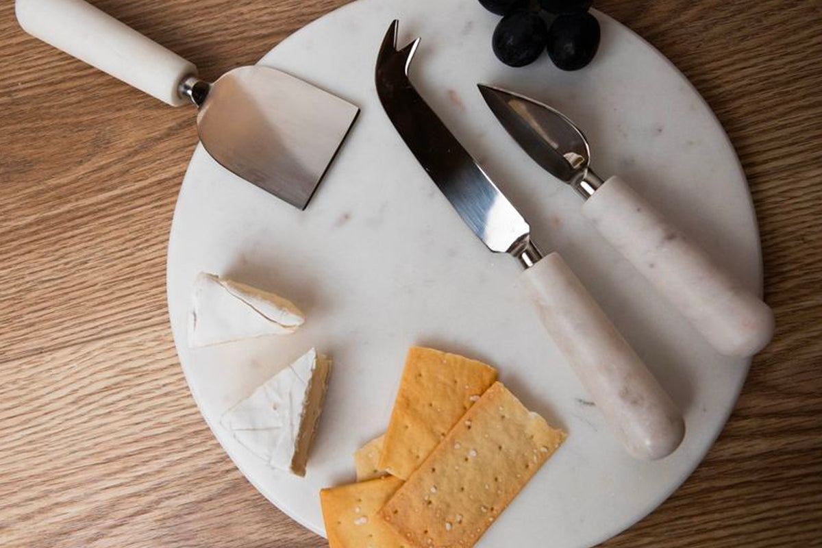 White marble round board with three stainless steel tools: cheese knife, spreader, and fork with white handles.