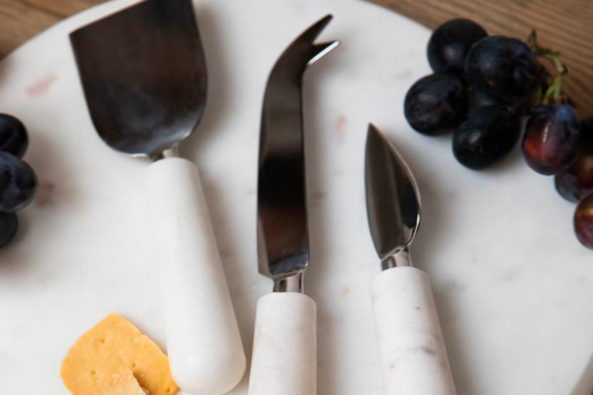Three stainless steel cheese tools with white marble handles displayed on a marble board with grapes and cheese.