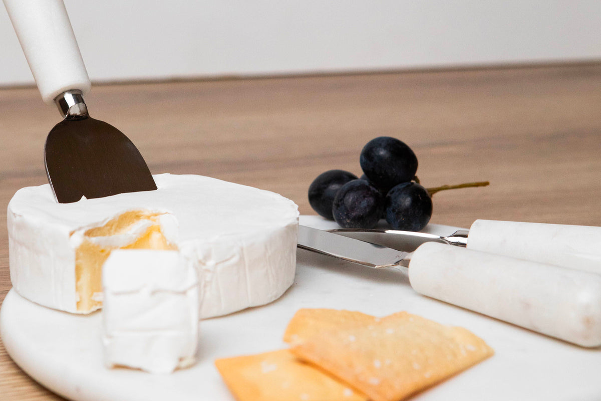 Three marble cheese tools with white handles displayed on a marble board with cheese and grapes.