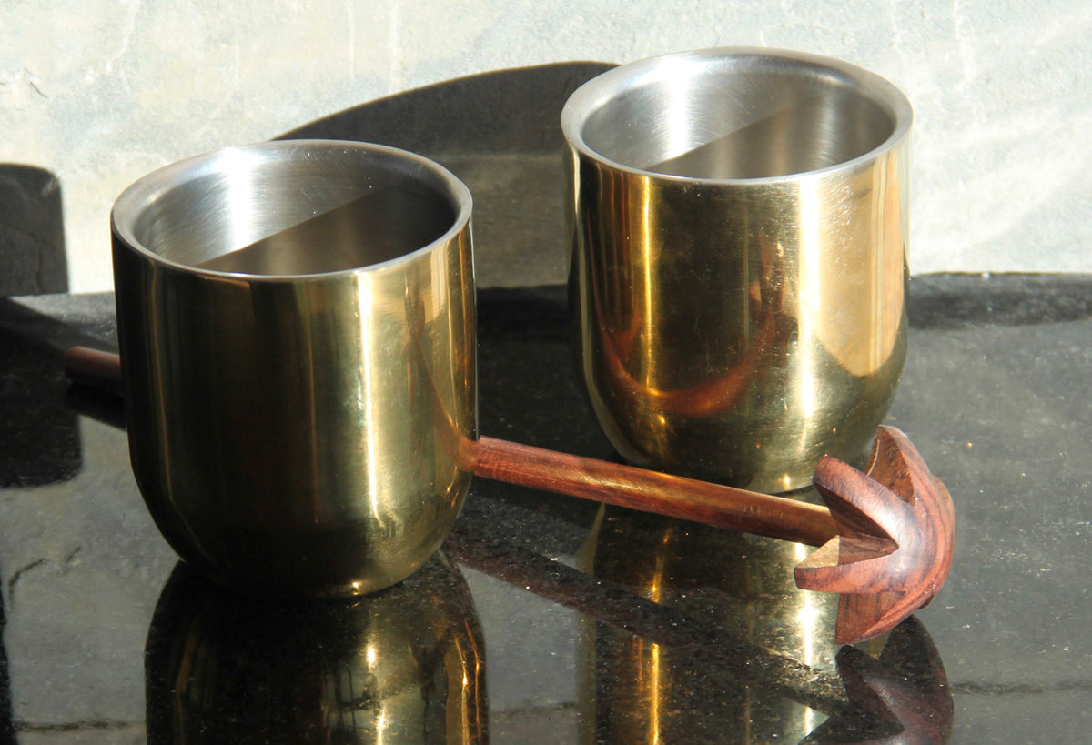 Two brass cups with wooden handle whisk featuring splayed metal beaters for mixing lassi.