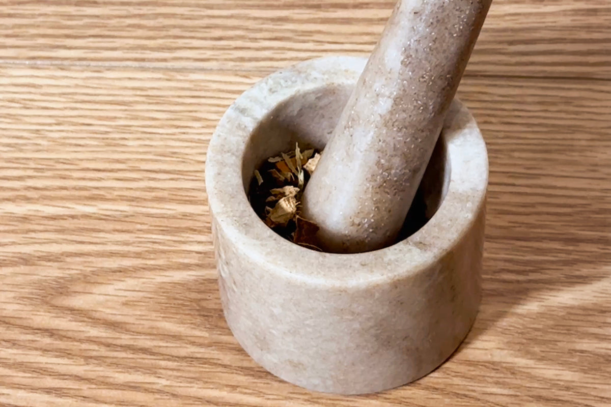 Cream-colored marble mortar with matching pestle containing dried cardamom pods on wooden surface.