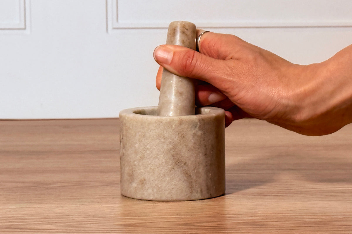 Hand holding wooden pestle in small round marble mortar and pestle set on wooden surface.