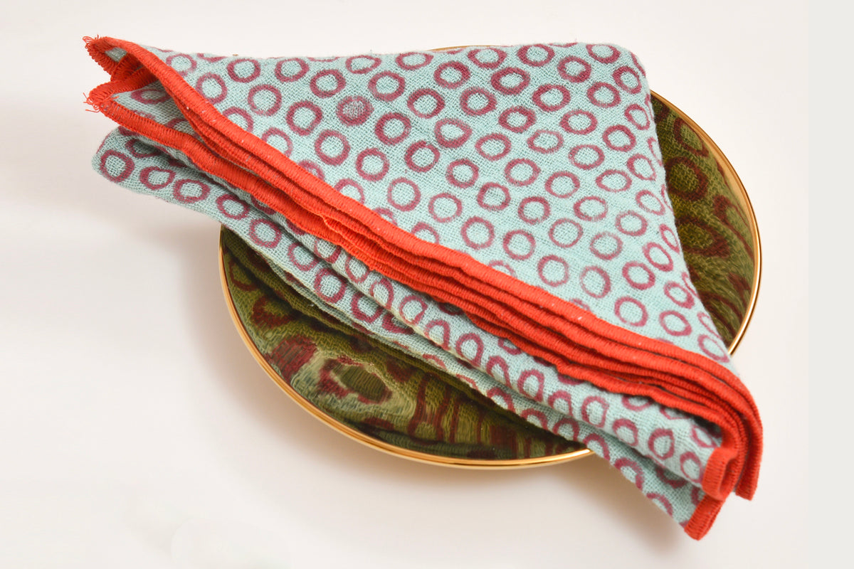 Folded napkins with red geometric block print pattern and red trim edges displayed on gold-rimmed plates.