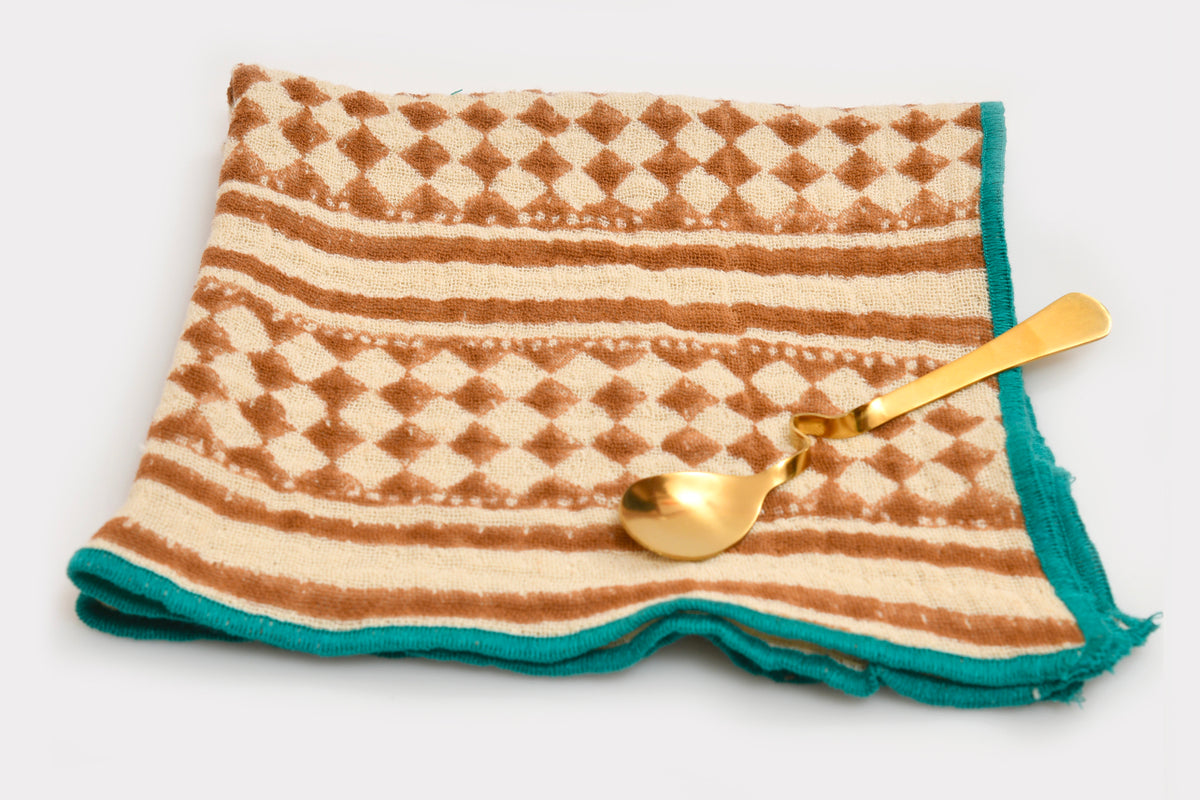 Cream and rust block print napkin with geometric diamond pattern, striped border, teal trim, and gold spoon.