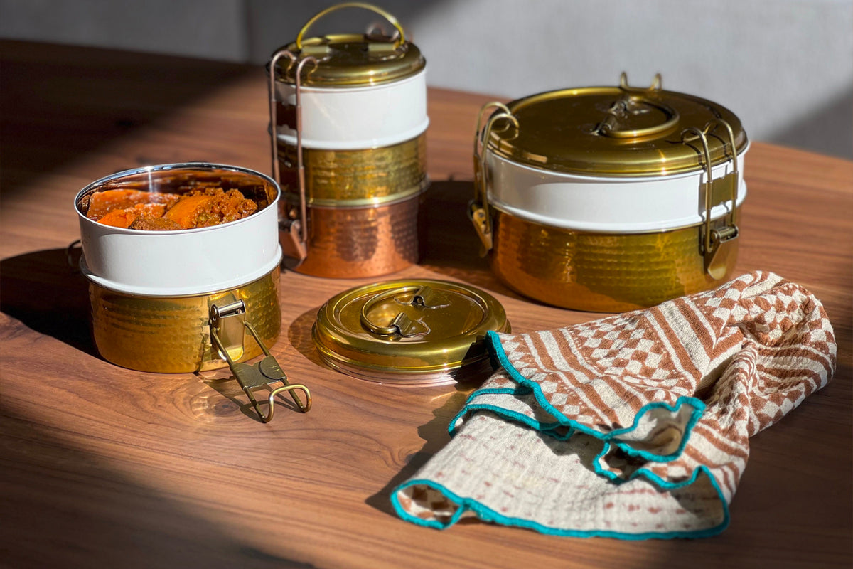 Four cream and rust block print napkins with turquoise trim displayed on wood surface next to brass containers.