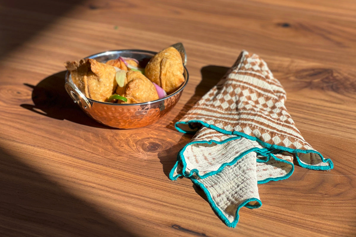 Folded napkin with geometric block print pattern and turquoise trim displayed on wooden surface next to bowl of fried snacks.