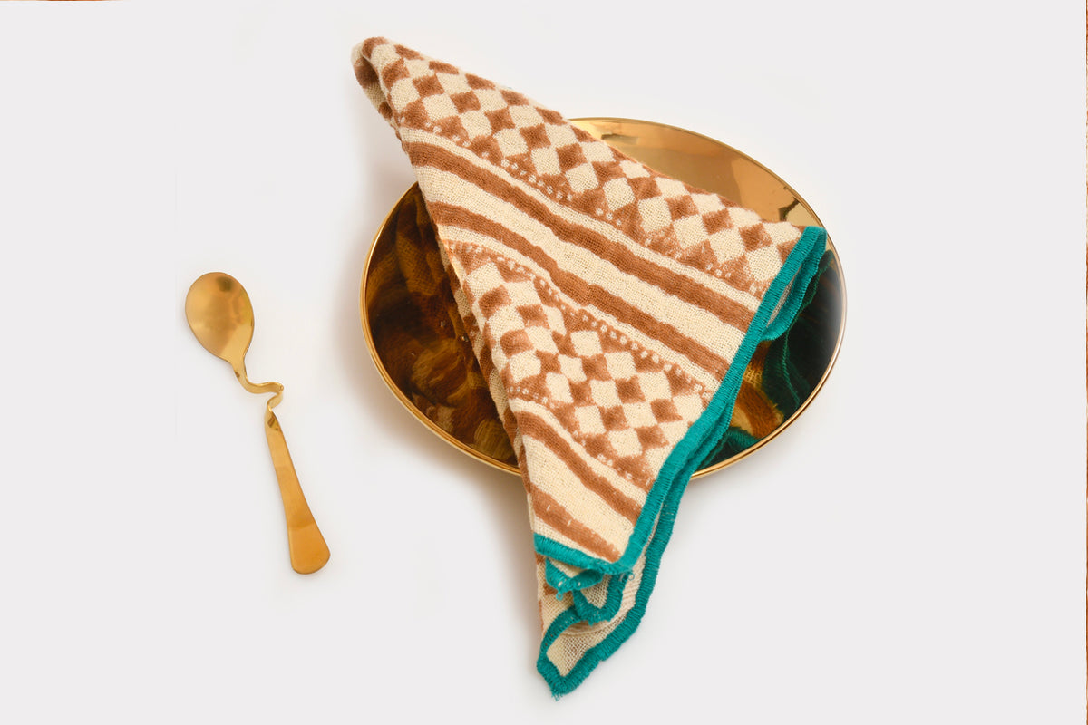 Cream napkin with terracotta geometric block print and teal border, draped on gold-rimmed plate with gold spoon.