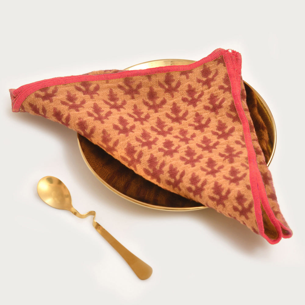 Tan napkin with burgundy geometric block print pattern and pink trim edges, displayed on wooden plate with gold spoon.