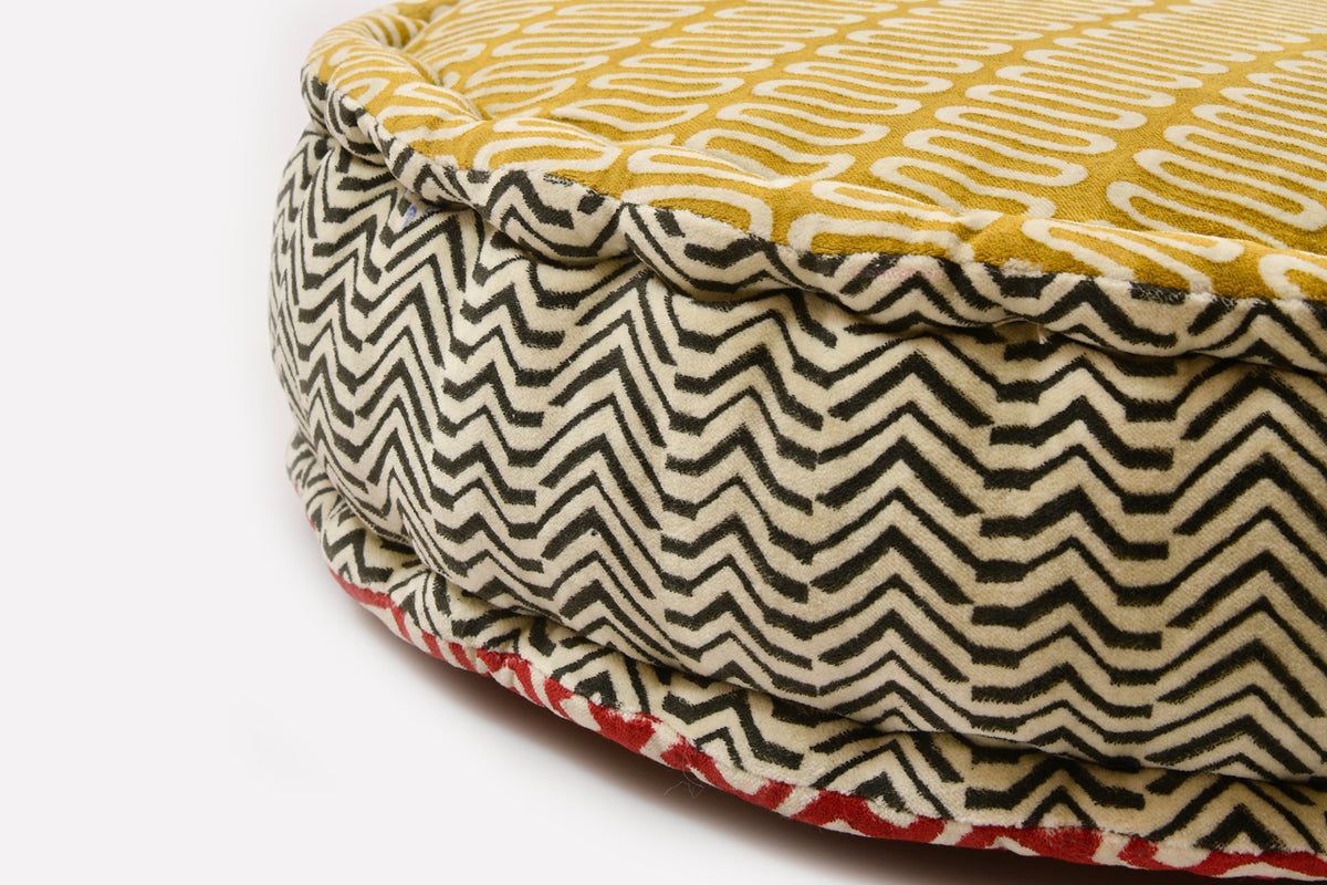 Round cushion with gold geometric block print on top and black/cream zigzag pattern on sides with red accent trim.