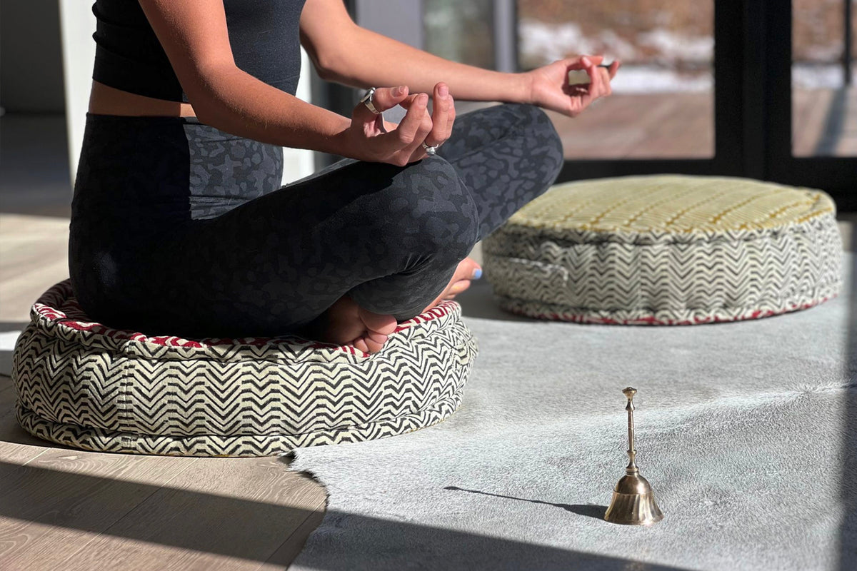 Person sitting cross-legged on round block print meditation cushion with geometric pattern in natural light.