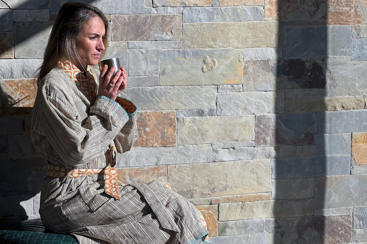 Woman wearing cream and brown block print reversible robe with patterned belt, seated against stone wall.