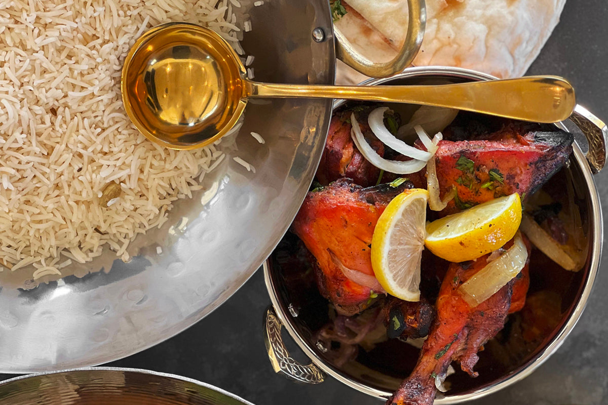 Gold-toned stainless steel serving spoon with flat bowl and long handle, displayed above cooked rice and spiced meat dishes.