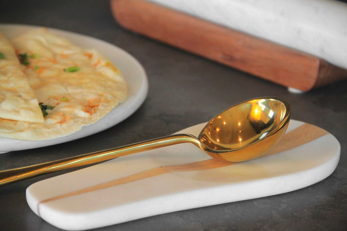 Gold-toned serving spoon with elongated bowl resting on a white ceramic spoon rest.