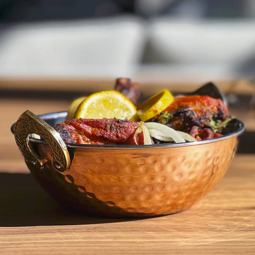 Hammered copper bowl with decorative handles containing cooked meat, lemon slices, and vegetables on wooden surface.