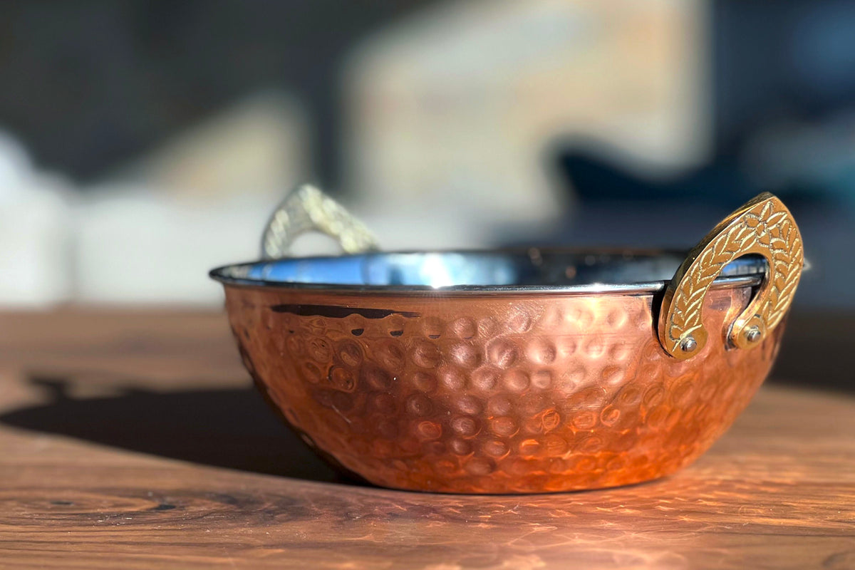 Copper handi serving bowl with hammered surface texture, blue interior, and brass handles.