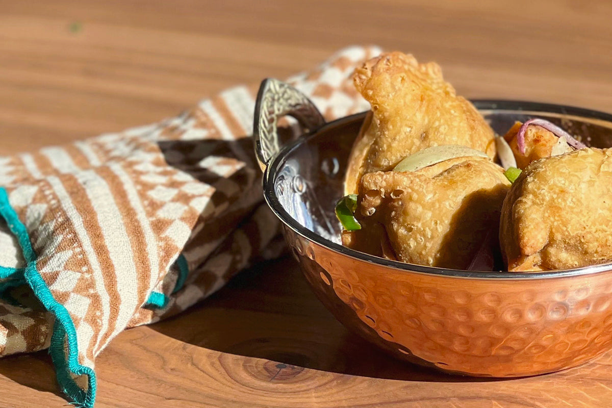 Hammered copper bowl with dark rim containing fried food and a serving spoon, placed on wooden surface with striped cloth.