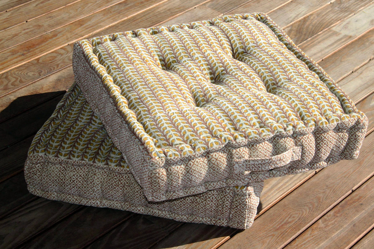 Square meditation cushion with geometric block print pattern in yellow, green, and gray on wooden floor.