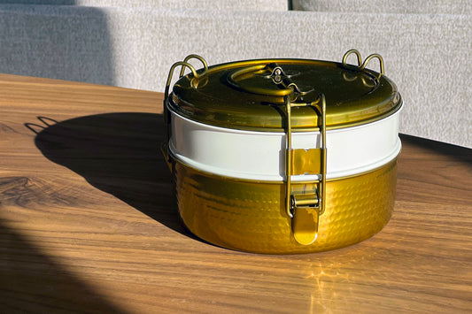 Stainless steel tiffin lunch box with gold hammered finish, white band, and metal latch closure on wooden surface.