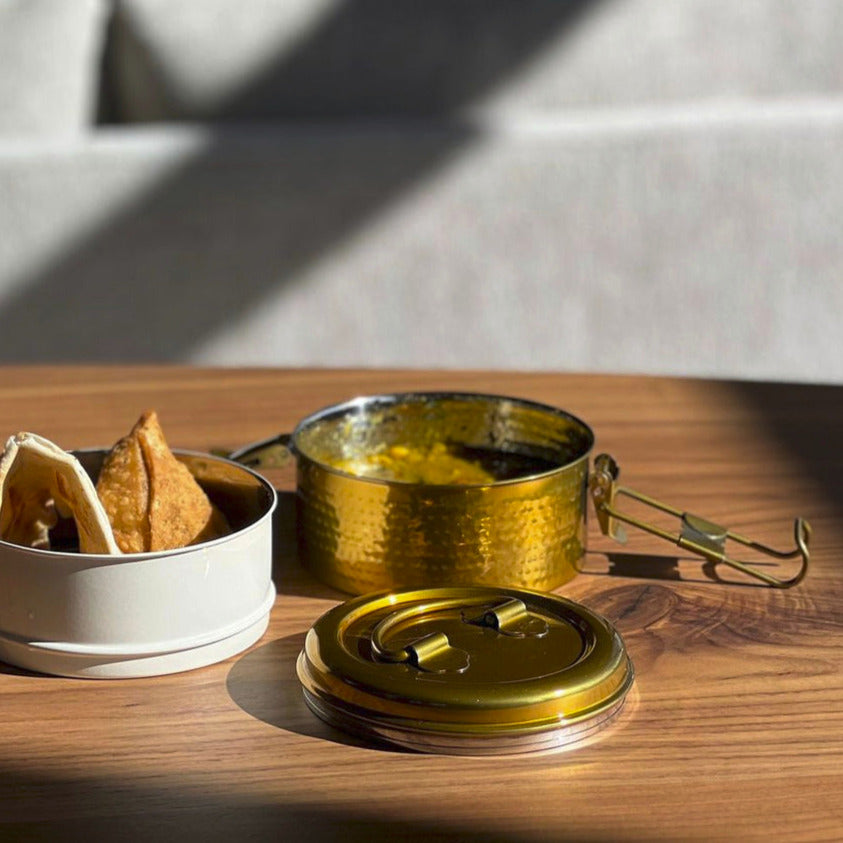 Stainless steel tiffin lunch box with brass finish, white container, and handles displayed on wooden surface.