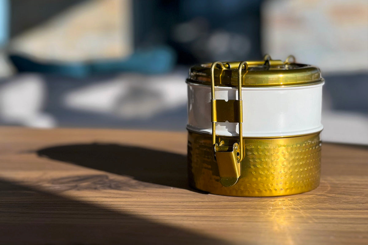 Stainless steel tiffin lunch box with gold hammered base, white band, and brass latch closure.