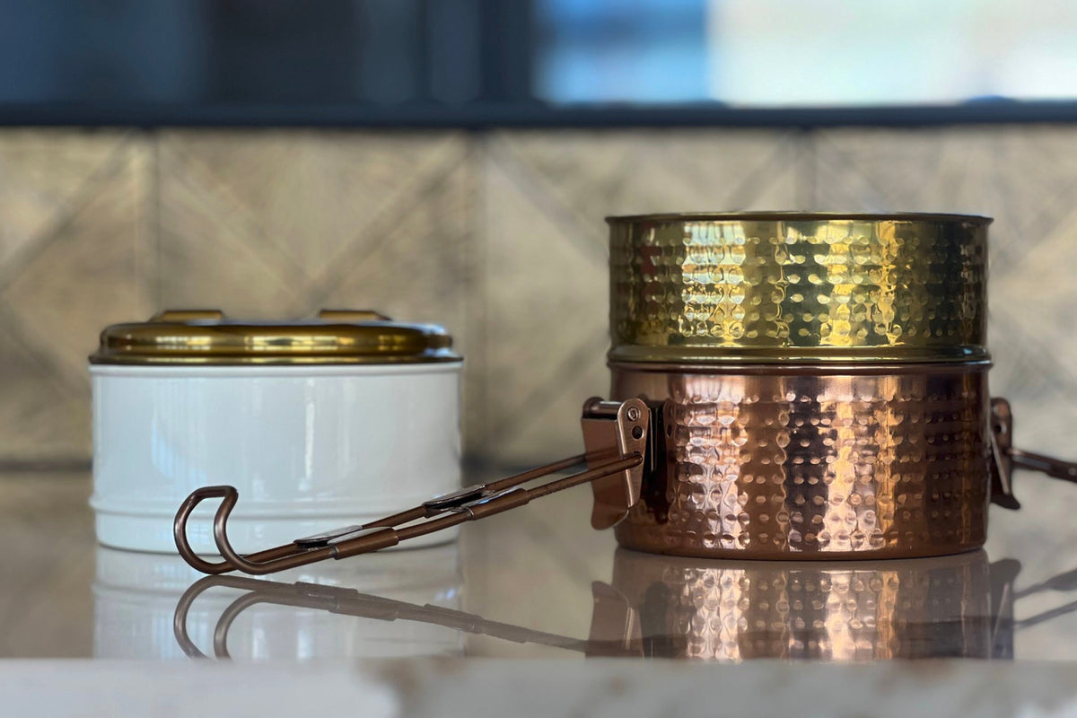 Two-tier tiffin lunch box with white ceramic bottom container and hammered copper top tier, connected by metal handle.