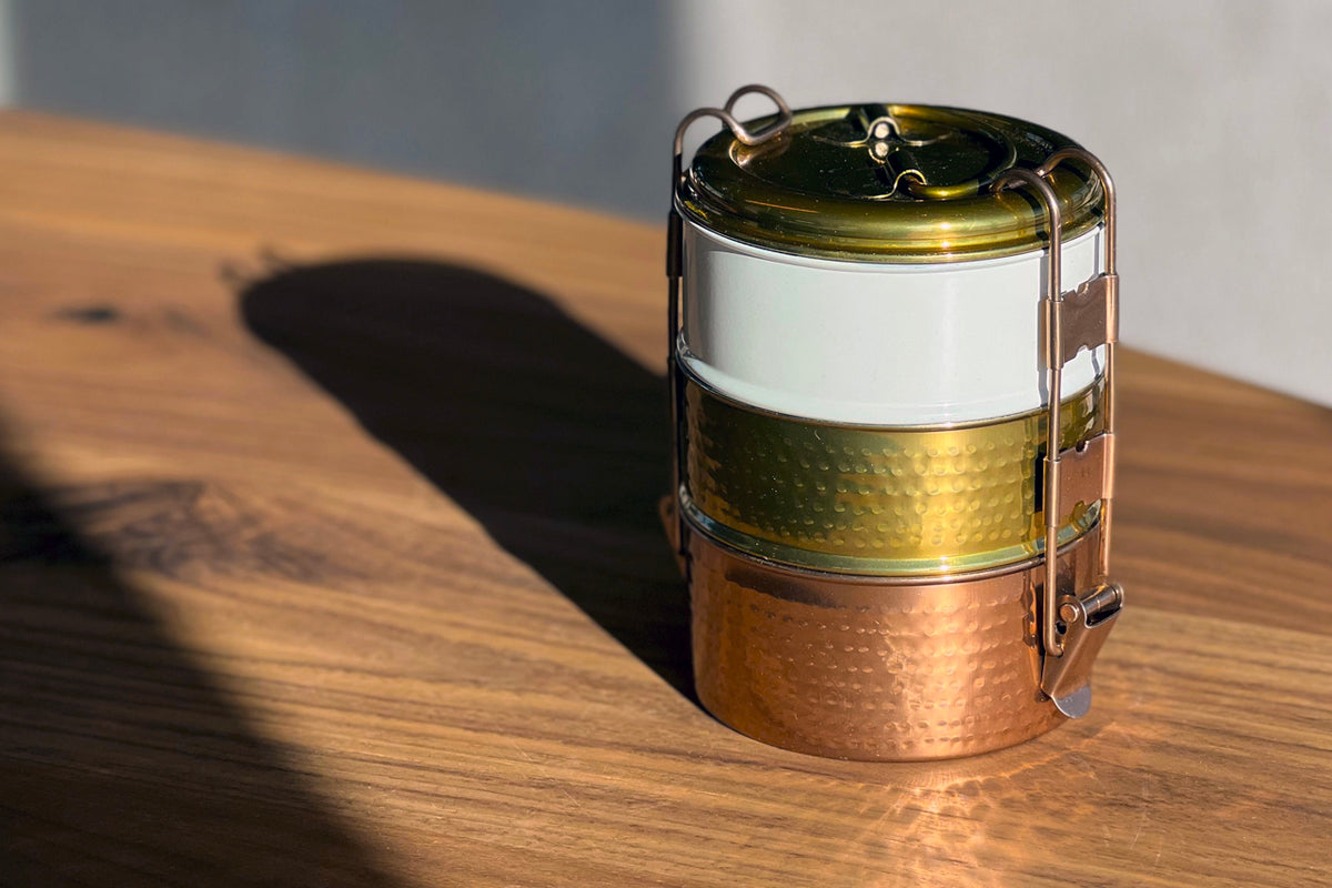 Three-tier stainless steel tiffin lunch box with brass and copper finish, carrying handles, and white band separator on woode