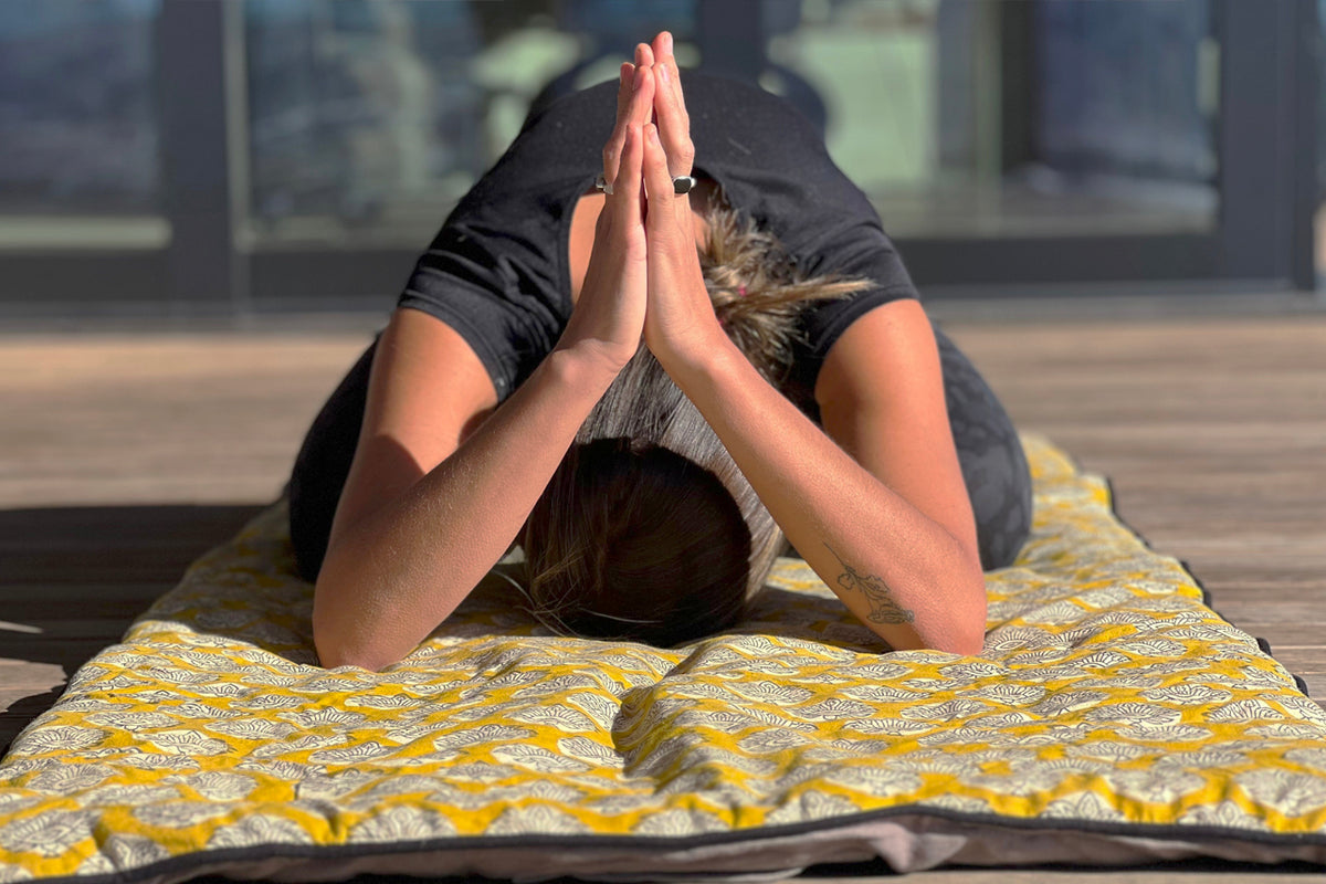 Person in forward fold pose on yellow block print yoga mat with geometric pattern.