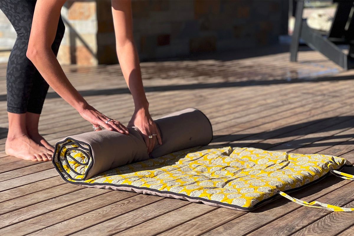 Person rolling a gray yoga mat with yellow block print towel laid out on wooden deck.