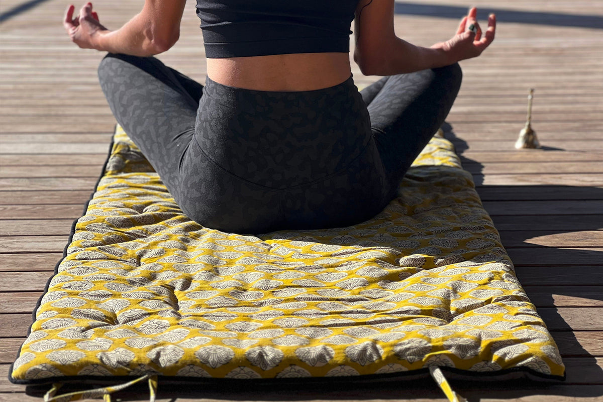 Person in meditation pose on yellow and white block print yoga mat outdoors on wooden deck.