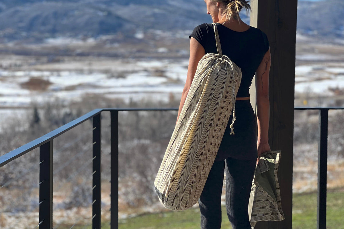 Woman carrying cream-colored yoga mat in woven carrying case with matching towel bag over shoulder.