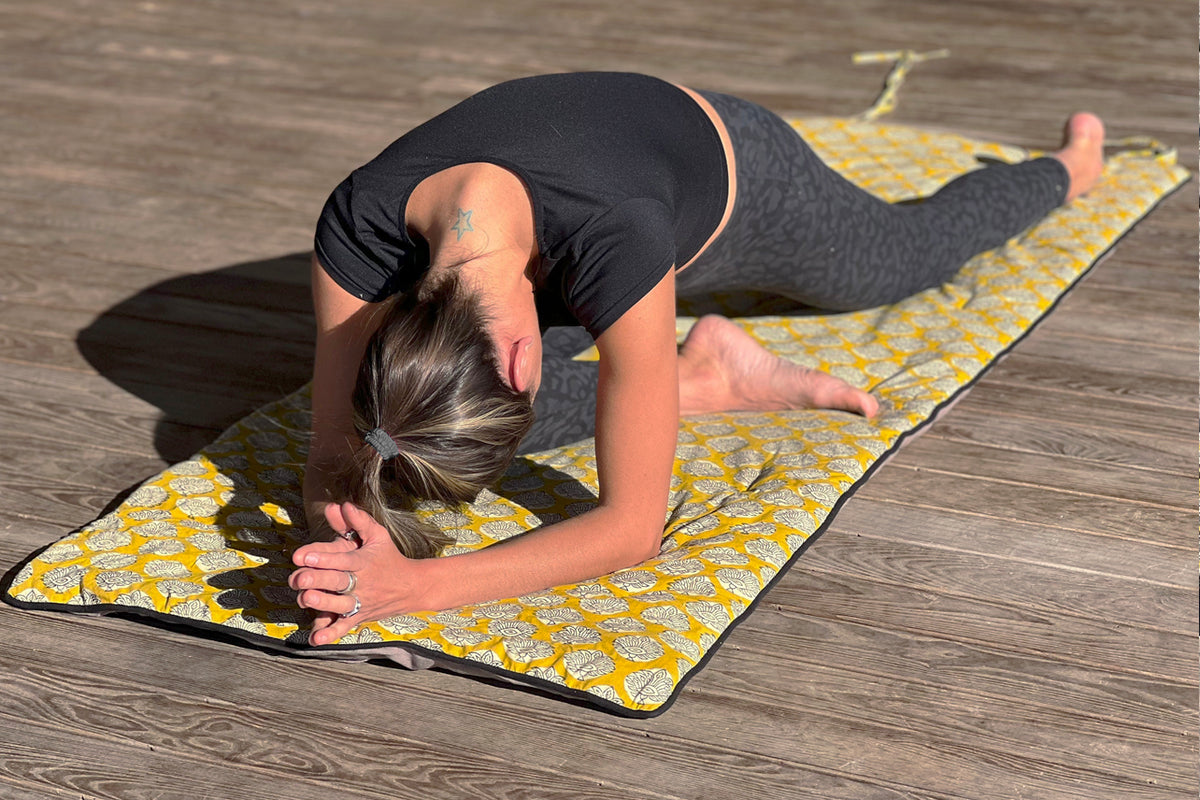 Person in forward bend pose on yellow block print yoga mat with matching towel on wooden floor.