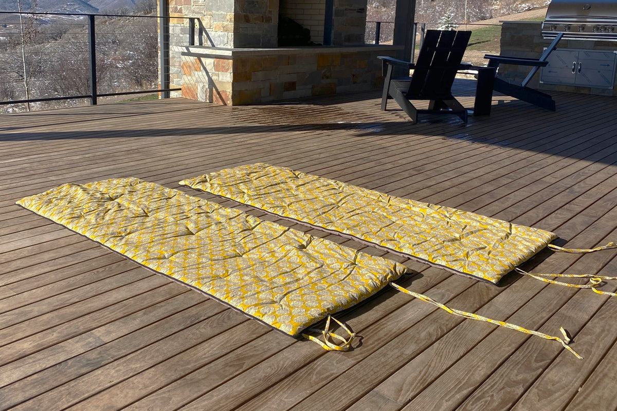Two yellow block print yoga mats with matching towels rolled and secured with straps on a wooden deck.