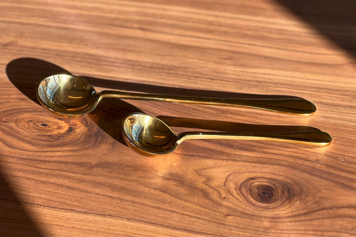 Two gold-toned serving spoons with elongated bowls and slender handles on a wooden surface.