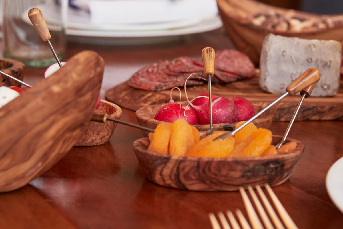 Wooden olivewood serving set with small bowls, appetizer picks with wooden handles, and cured meats arranged on a table.