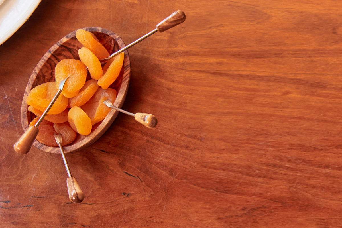Wooden olivewood serving dish with three metal pick utensils holding dried apricots.