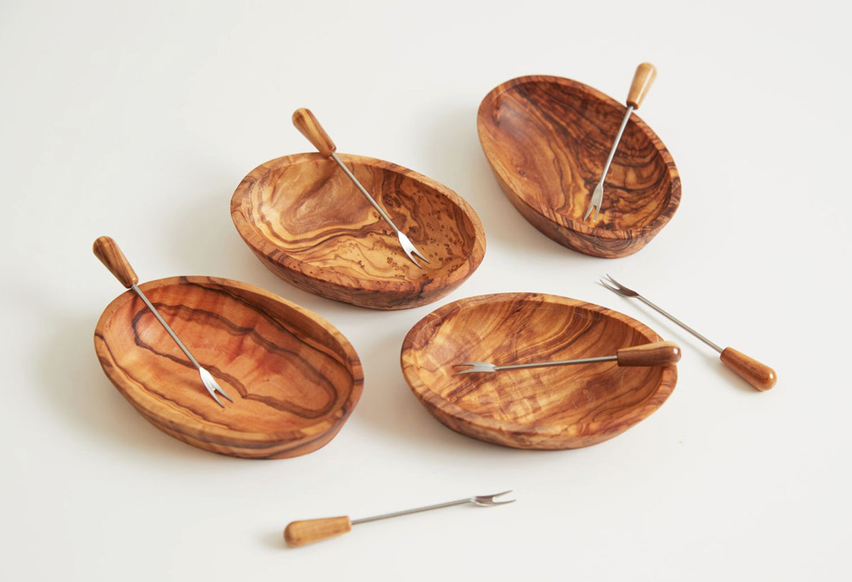 Four olive wood bowls with metal serving spoons and two forks with wooden handles on white background.