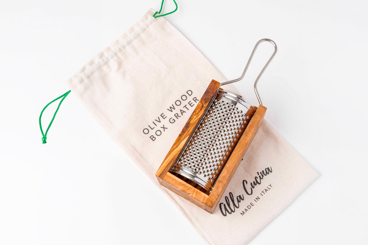Box grater with olivewood frame and metal grating surface, shown with cream cotton storage pouch featuring green drawstring.