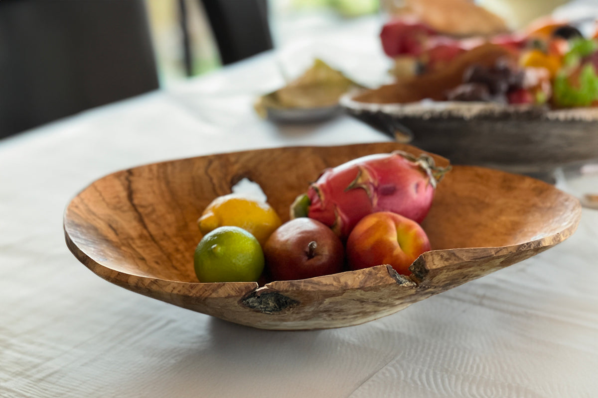 Wooden bowl with organic grain pattern holding fresh fruit including apples, limes, and pomegranate on white table.