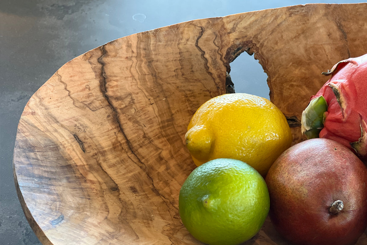 Olivewood bowl with organic grain patterns and cutout handles holding lemon, lime, pomegranate and strawberry.