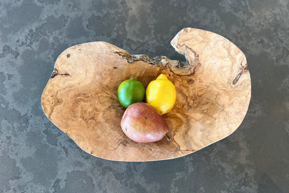 Organic-shaped wooden bowl with lime, lemon, and apple inside, displayed from above on gray surface.