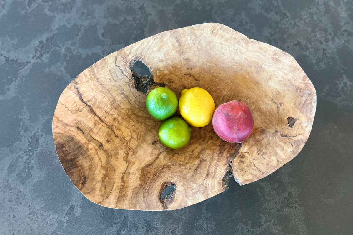 Natural wood bowl with irregular edges holding green apples, yellow lemon, and red apple on gray surface.