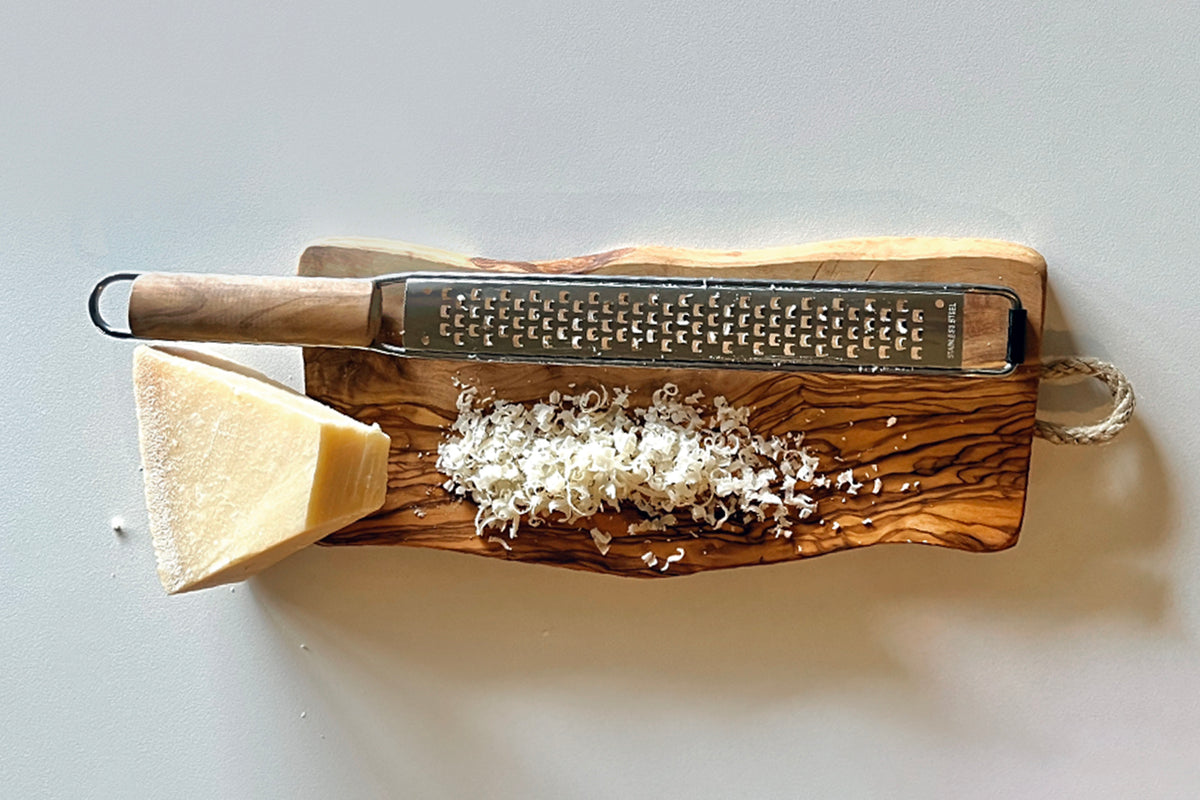 Olivewood paddle grater with metal grating surface, wooden handle, and shredded cheese on wooden board.