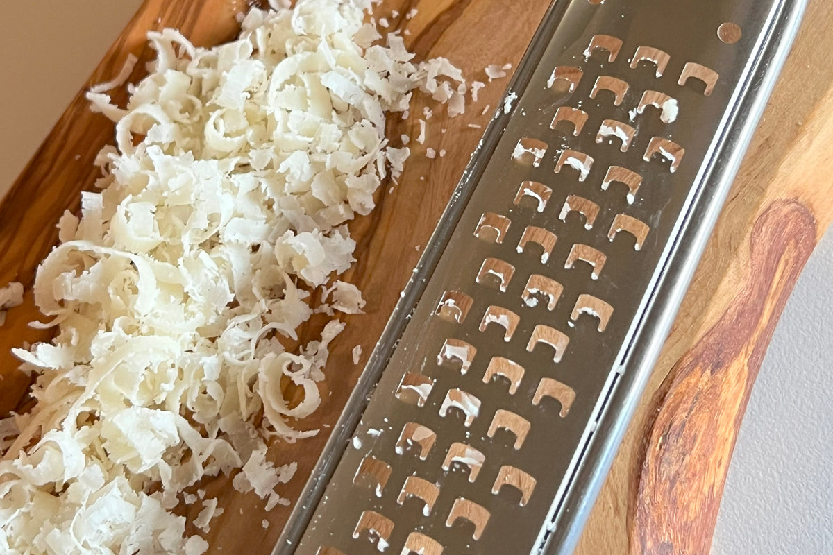 Olivewood mandoline grater with metal grating surface, shown grating cheese onto wooden board.