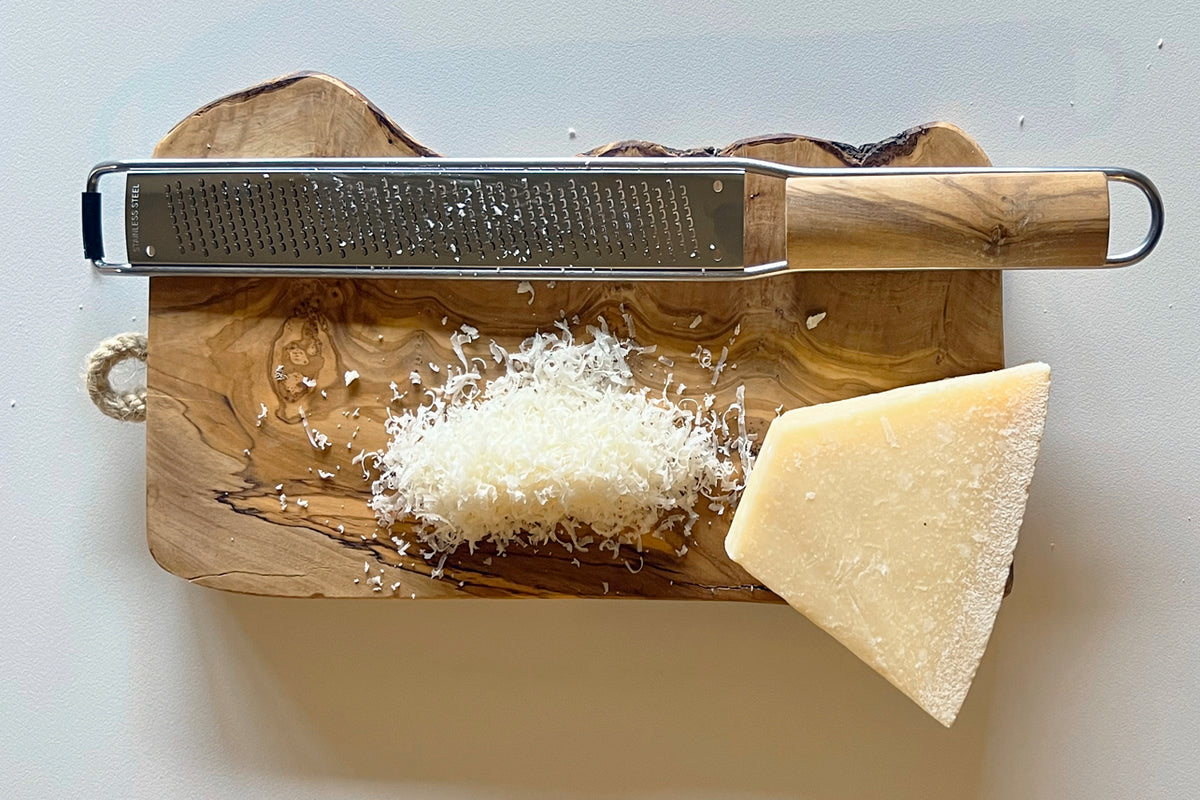 Olivewood paddle grater with metal grating surface and wooden handle, displayed on wooden board with grated cheese and cheese