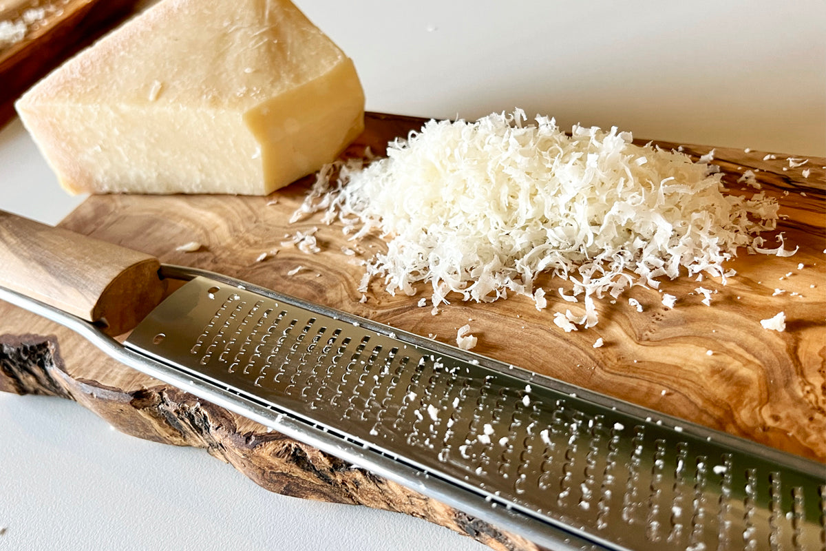 Olivewood paddle grater with metal grating surface next to grated cheese and a wedge of hard cheese on a wooden board.